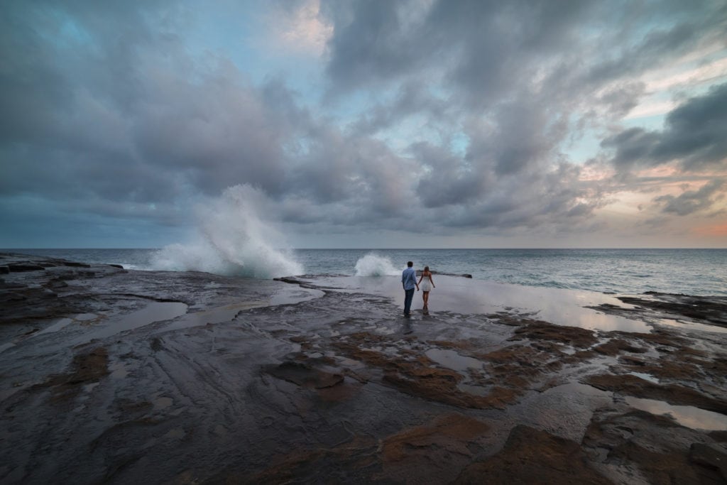 A couple enjoying a Hawaiian sunset while waves crash against the rocks at Lanai Lookout during their elopement.