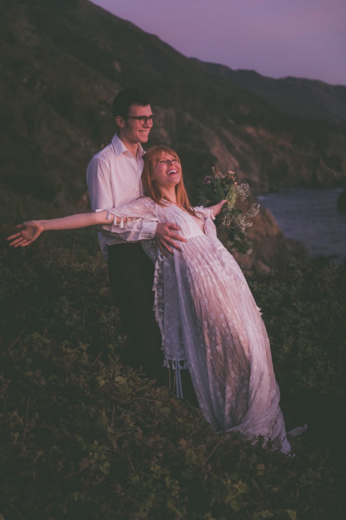 A groom holds the bride while she goofs off during sunset in Big Sur.